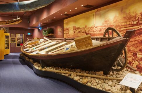 A large York boat is nestled amongst larger rocks with artifact cases inside made to look like fur bales but featuring artifacts related to freighting and transportation. The walls of the gallery are a deep burgundy and there is a mural behind the boat depicting scenes from HBC’s prominent trading post at Norway House.