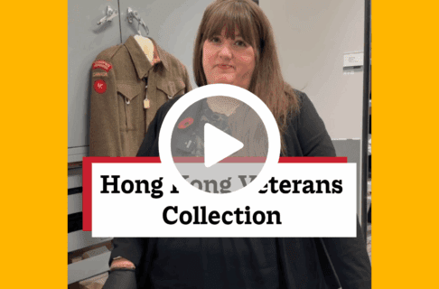 A screenshot of a video, an individual stands inside museum collection storage, with a Hong Kong uniform hanging behind. There's a play button over the screenshot and overlaid text reads, "Hong Kong Veterans Collections".