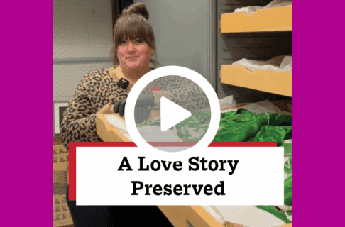 A screenshot of a video, an individual stands inside museum collection storage, next to an open drawer displaying green silk pyjamas. There's a play button over the screenshot and overlaid text reads, "A Love Story Preserved".