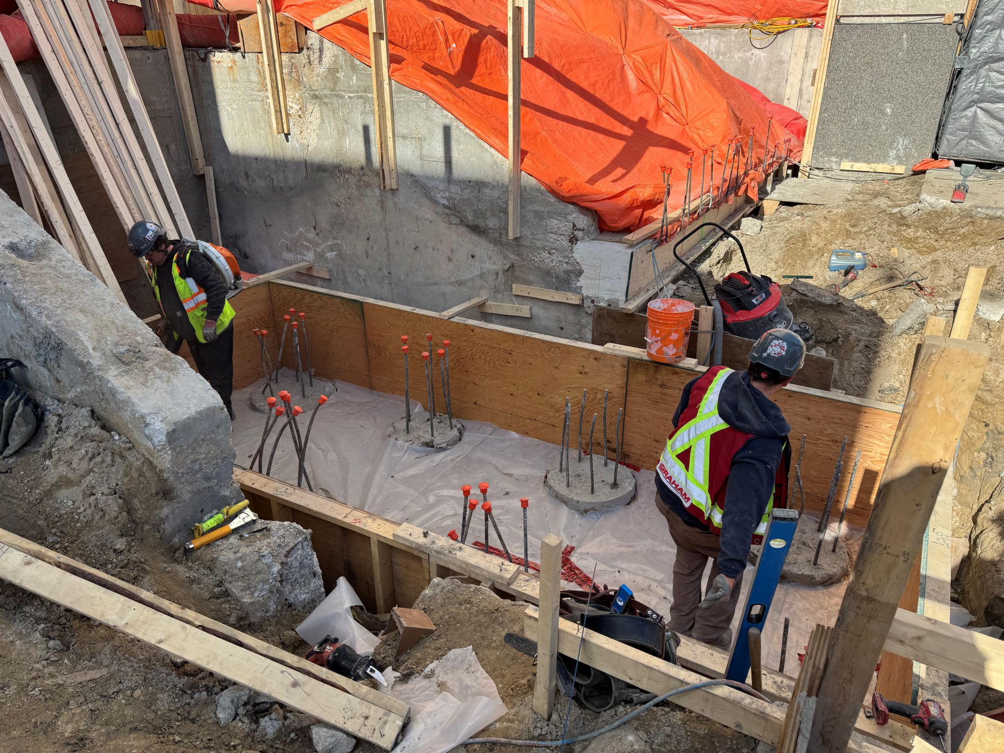 Two individuals wearing high vis vests over their work wear in a pit of a construction site. In the upper portion of the frame part of an orange tarp can be seen covering an area.