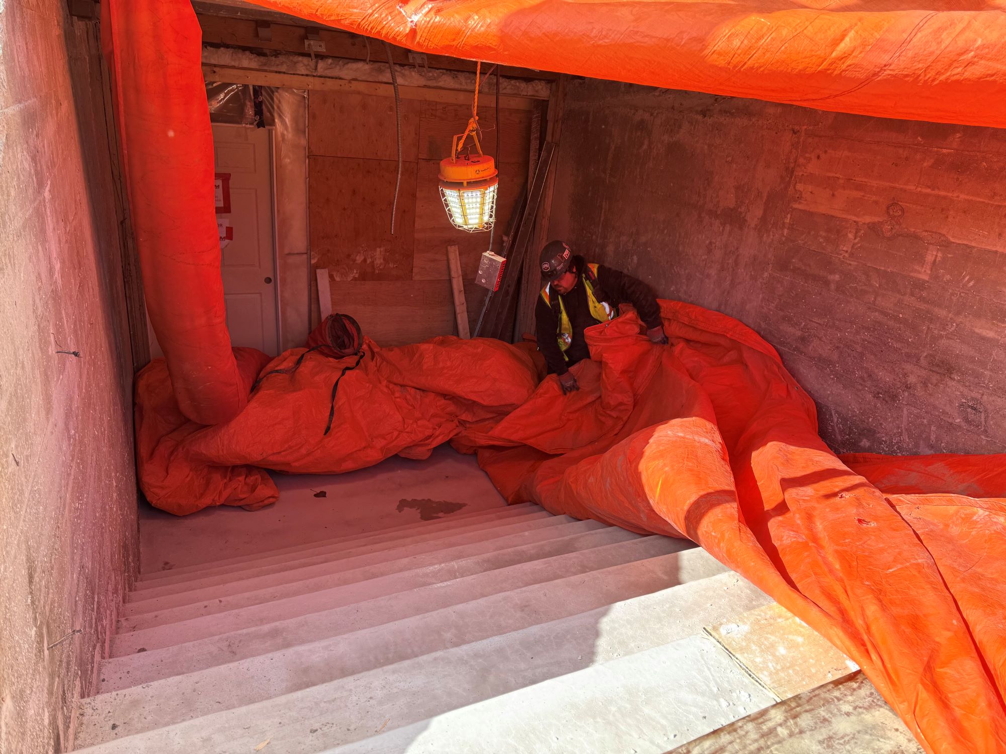 A construction worker holds back an orange tarp covering a cement stairwell leading down to a barricaded door.
