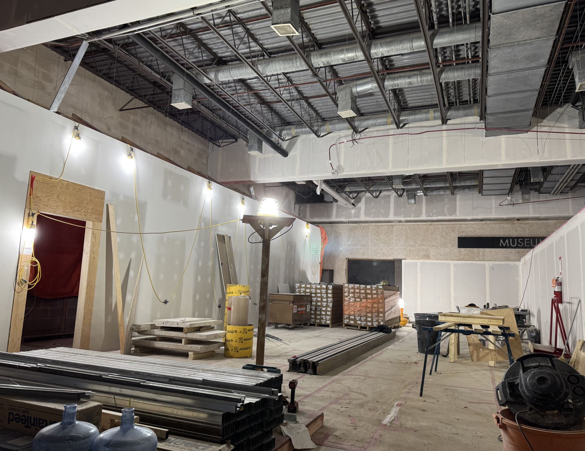 Looking into an interior construction site, with materials stacked about. The ceiling is uncovered, showing the pipes and vents, with all light fixtures removed.