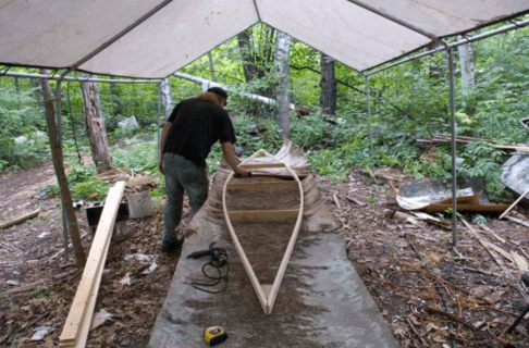 The wooden frame of a canoe laid on a sheet of birch bark beneath and open-sided tent in a wooden area.
