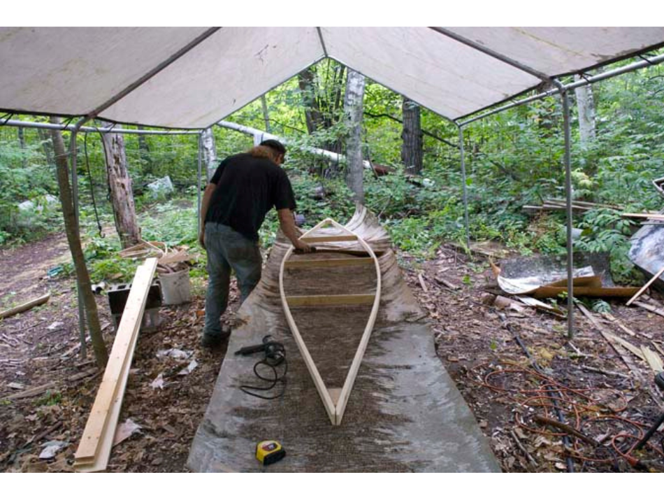 The wooden frame of a canoe laid on a sheet of birch bark beneath and open-sided tent in a wooden area.