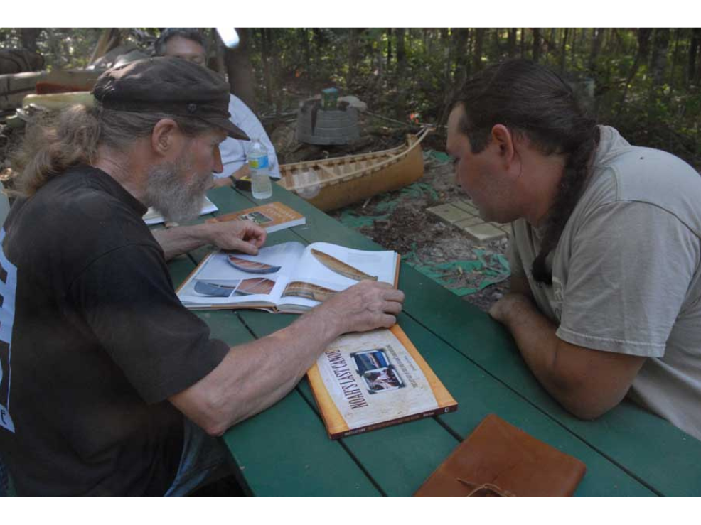 Two individuals with heads close together seated on either side of a picnic table looking at a book showing styles of canoes.