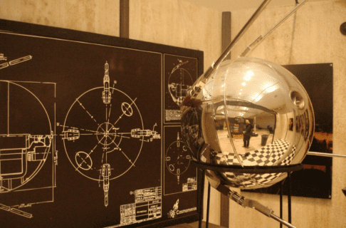 A reflective metal orb on a stand in front of a chalkboard like sign with blueprints on it.