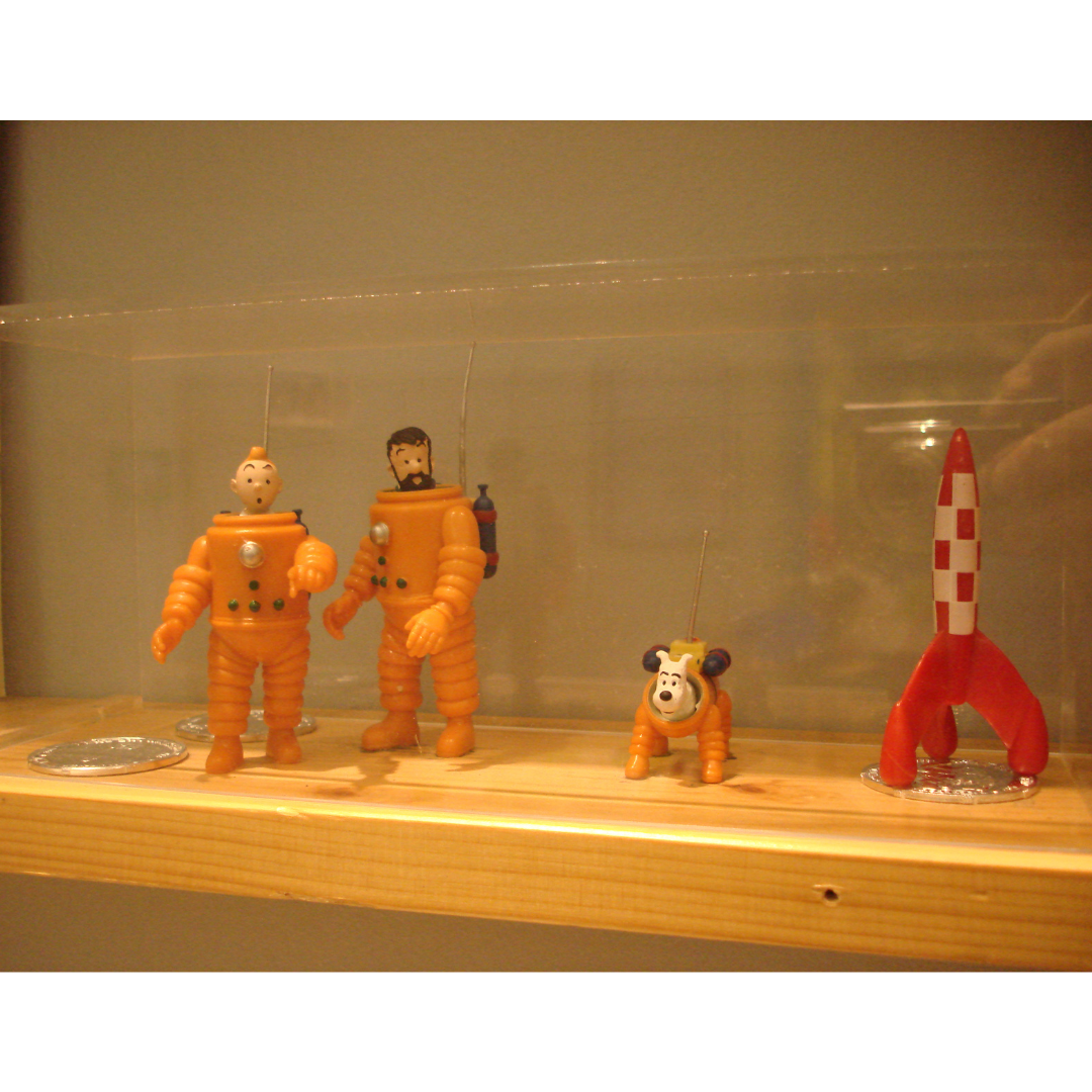 Figures of Tintin, Captain Haddock, and Snowy the dog in orange space suits next to a red rocket.