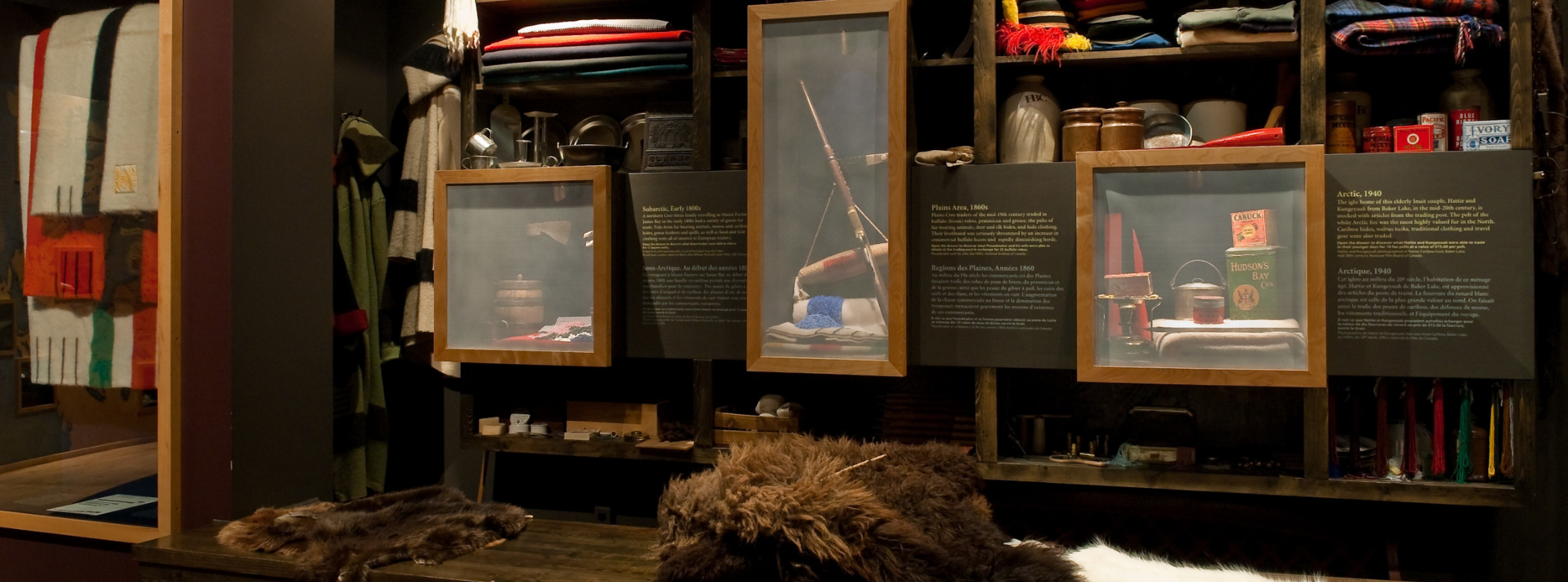 A long wooden countertop with a brown beaver pelt, a folded up brown bison pelt, and a white arctic fox pelt on the top. Behind the counter is a mock trading post, with shelves lined with trade goods like coloured cloth and fabric, metal dishes, ceramic crocks, and smaller items for sewing. There are three lit-up cases that feature artifacts as examples of items that individuals could get at a trading post.