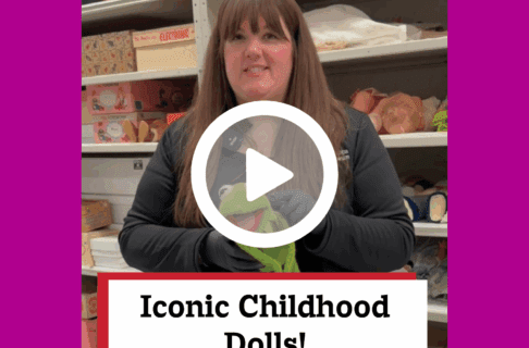 A screenshot of a video, an individual stands inside museum collection storage holding a stuffed Kermit the frog toy. There's a play button over the screenshot and overlaid text reads, "Iconic Childhood Dolls!".