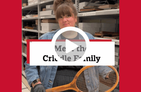 A screenshot of a video, an individual stands in museum collections storage holding a vintage tennis racket. There's a play button over the screenshot and overlaid text reads, "Meet the Criddle Family".