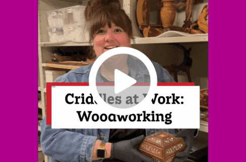 A screenshot of a video, an individual stands in museum collections storage holding a wooden box with mother of pearl inlay. There's a play button over the screenshot and overlaid text reads, "Criddles at Work: Woodworking".