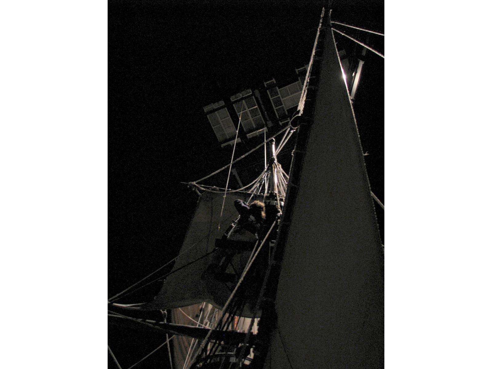 View up into the ratlines and sails of a wooden sailing vessel in an indoor gallery. In the darkness above, the base of a temporary platform can be seen