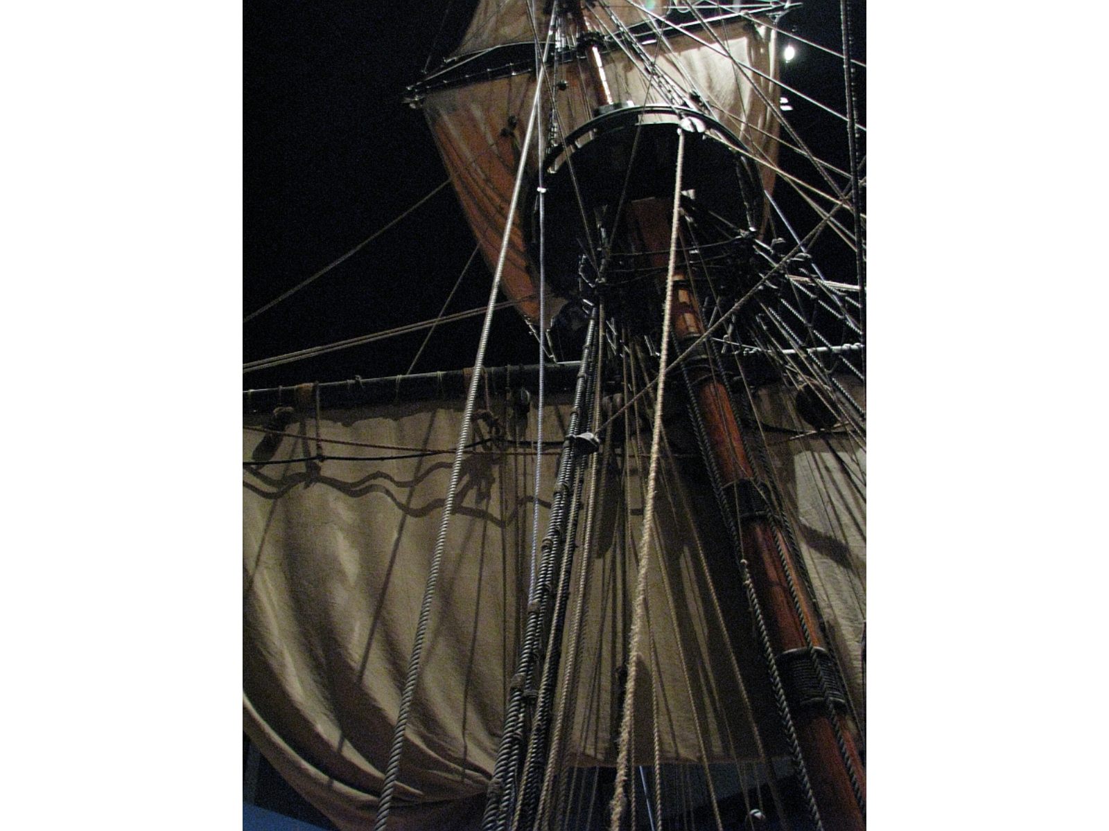 View up into the ratlines and sails of a wooden sailing vessel in an indoor gallery.