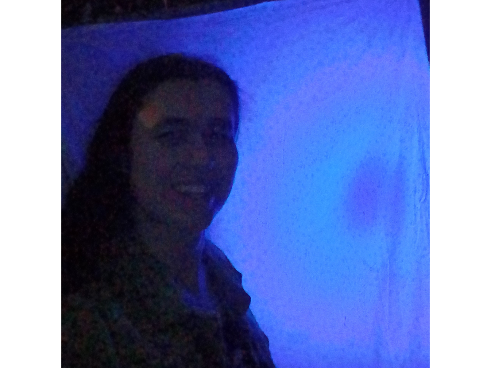 A nighttime selfie of a smiling individual standing in front of a hanging sheet that is lit blue by a UV flashlight.