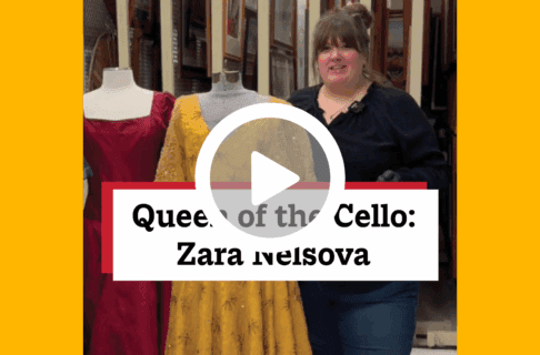 A screenshot of a video, an individual stands in collection storage next to a red gown and yellow gown on dress forms. There's a play button over the screenshot and overlaid text reads, "Queen of the Cello: Zara Nelsova".