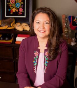A formal headshot of a smiling woman wearing a plum-coloured blazer with beadwork detailing on the lapels.