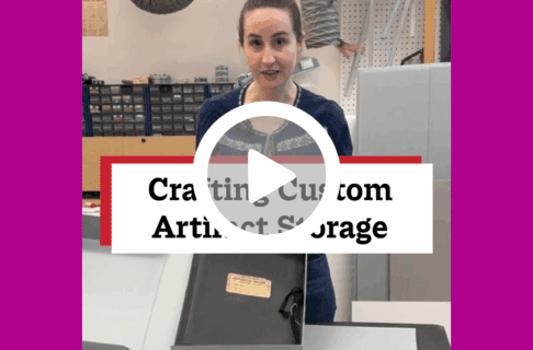 A screenshot of a video, an individual stands in front of a table in a museum lab. holding up a open clamshell box with a scrapbook in it There's a play button over the screenshot and overlaid text reads, "Crafting Custom Artifact Storage".