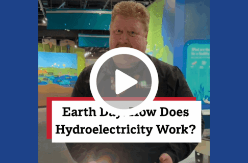 A screenshot of a video, an individual stands in the Science Gallery near the water table and Lake Winnipeg Shared Solutions exhibit. There's a play button over the screenshot and overlaid text reads, "Earth Day! How Does Hydroelectricity Work?".