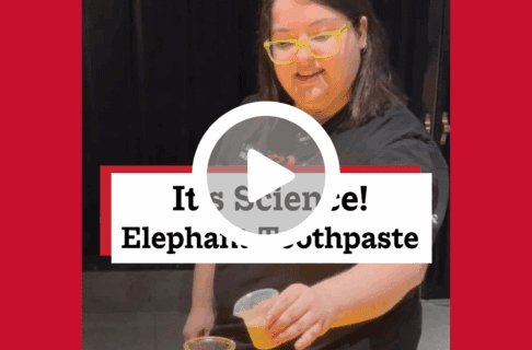 A screenshot of a video, an individual holds a small container to pour its contents into a vase below. There's a play button over the screenshot and overlaid text reads, "It's Science! Elephant Toothpaste".