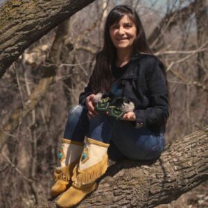 A woman sitting on a low-lying log while wearing mukluk boots and holding a pair of beaded and fur-cuffed moccasins.