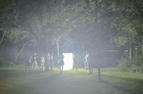 A bright rectangular light box, about the height of an average adult, being held up in a park at night. A small group of people all stand nearby.