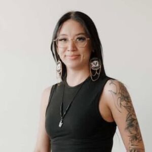 Formal headshot of a smiling individual with long dark hair and beaded earrings. Fine-line tattoos cover much of their left arm, as shown by their sleeveless shirt.