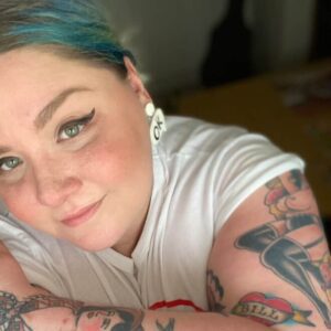 An informal selfie-style headshot of an individual with blue hair and thick winged eyeliner. Their left arm, partly visible in the image has several colourful tattoos.