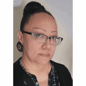 A selfie of a women with a neutral expression and dark hair in a top knot. She wears glasses and beaded earrings along with a beaded necklace.