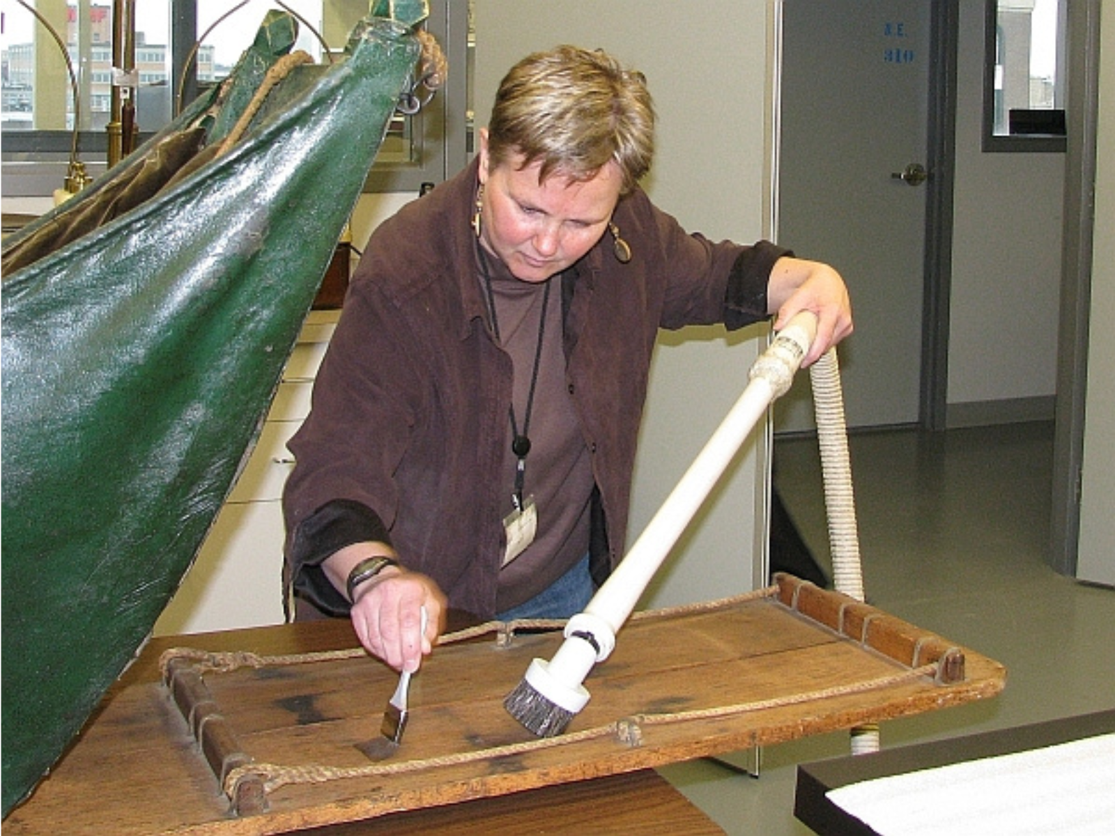 An individual holds up the nozzle and hose of a vacuum cleaner from out of frame, and with their other hand uses a small brush to brush dirt towards the mouth of the vacuum.