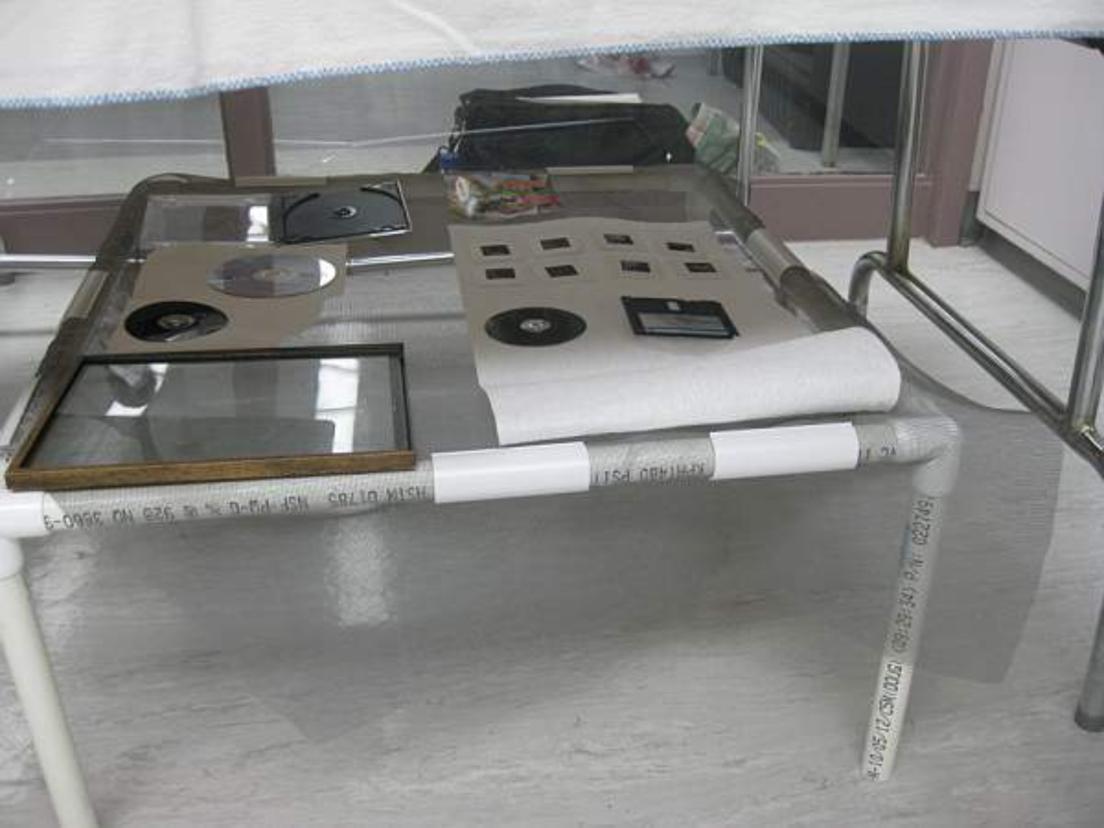 Objects like CDs, CD cases, floppy disks, and film slides laid on a small table made from pipe and netting.