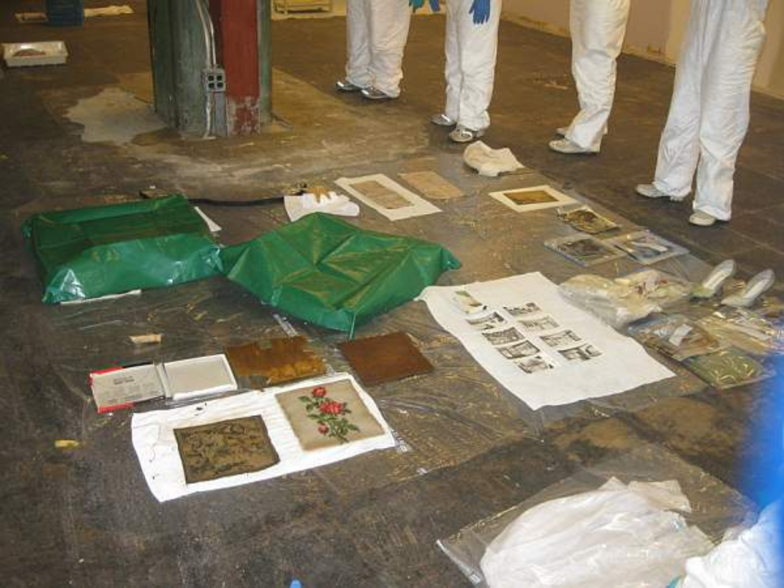 A series of painting an photographs laid out on paper towels on the floor. In the background the legs of four people wearing protective clothing can be seen.
