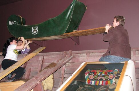 Three individuals at the ends of a green cariole sledge mounted on a wall.