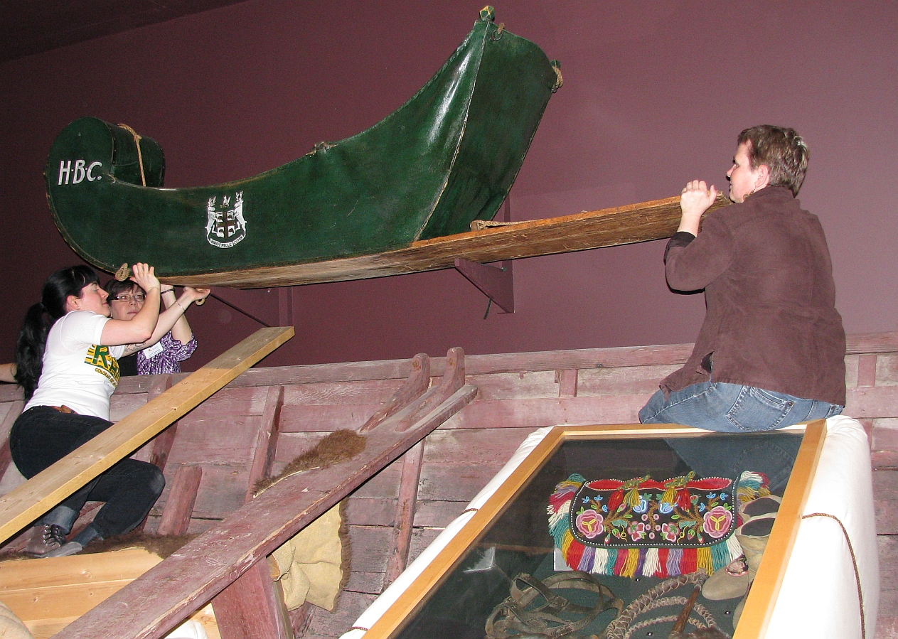 Three individuals at the ends of a green cariole sledge mounted on a wall.