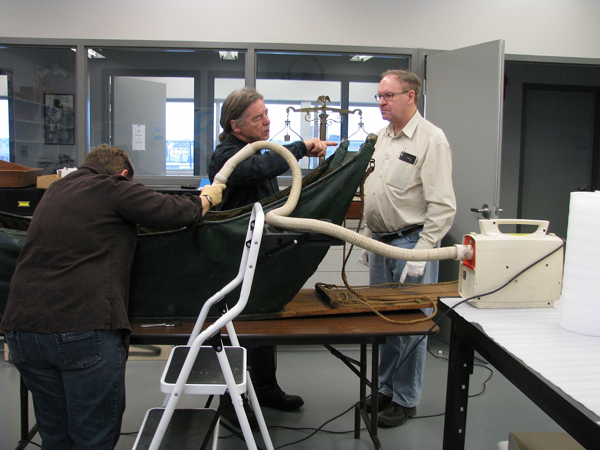Three individuals around a cariole sledge on a table in a museum lab. Two are in discussion, and the third is vacuuming the inside of the cariole.