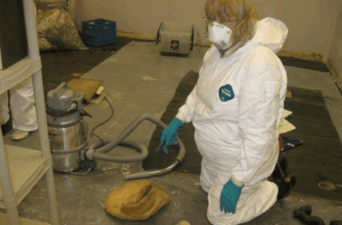 An individual wearing white protective clothing, blue rubber gloves, and a white face mask kneels on the ground next to a shop vac and an old and worn straw hat.