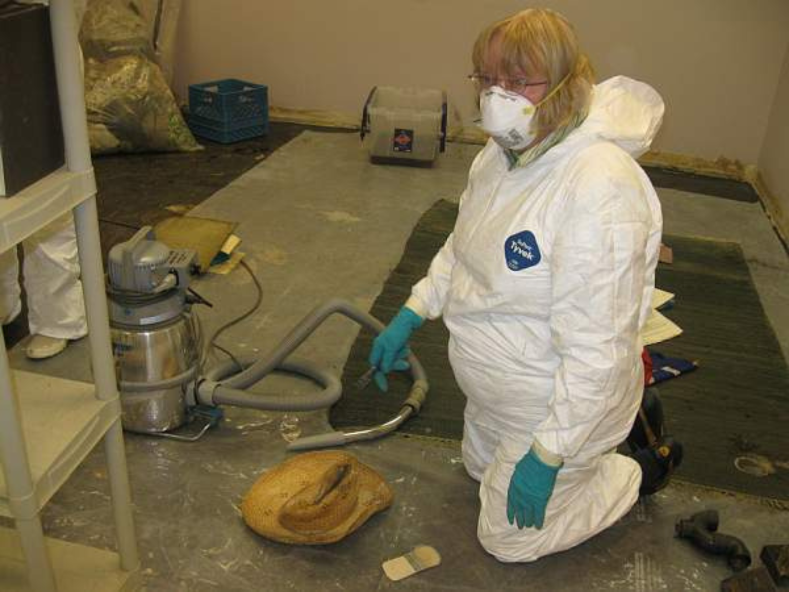 An individual wearing white protective clothing, blue rubber gloves, and a white face mask kneels on the ground next to a shop vac and an old and worn straw hat.