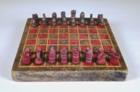 A rough, handmade chess set with red and platinum coloured pieces lined up in starting positions.