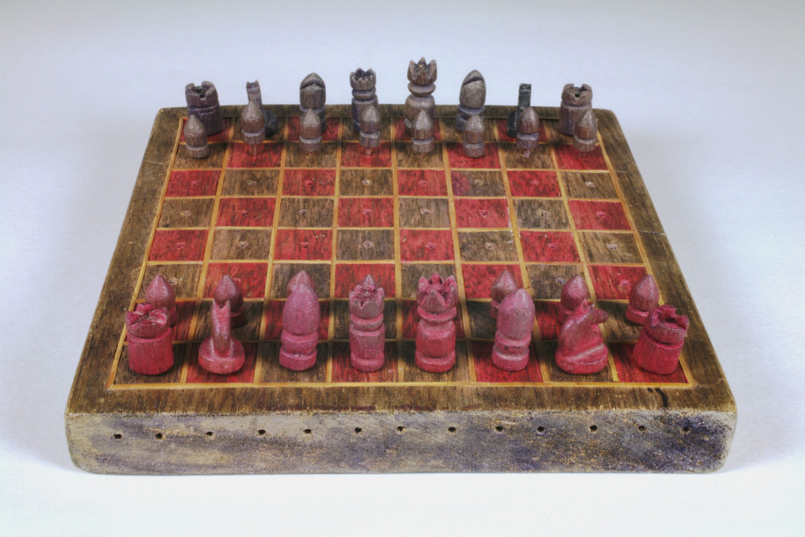 A rough, handmade chess set with red and platinum coloured pieces lined up in starting positions.