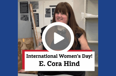 A screenshot of a video, an individual stands in front of a table in a museum lab. There's a play button over the screenshot and overlaid text reads, "International Women's Day! / E. Cora Hind".