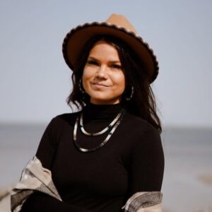 Photo of an individual wearing a dark shirt and a beaded necklace as well as a wide brimmed hat with a beaded brim.