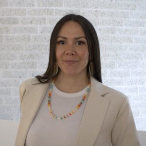 A headshot of an individual with long dark hair and a soft smile, wearing a light coloured top and blazer with a beaded necklace and earrings.