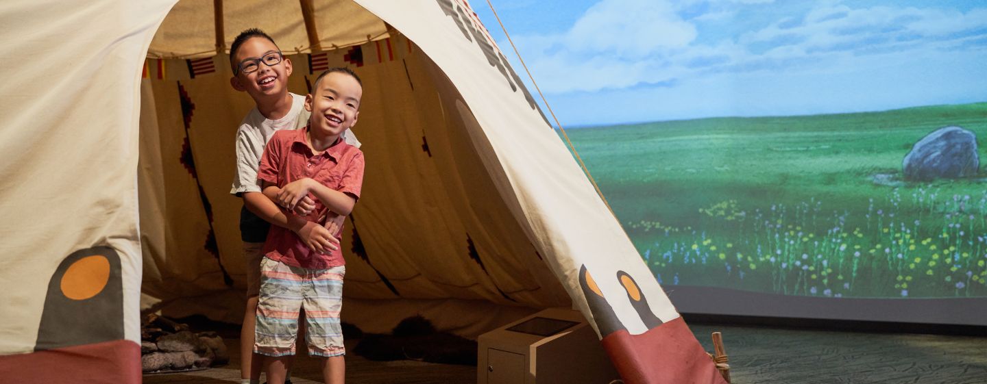 Two children laughing and hugging while standing in the opening of the tipi in the Manitoba Museum's Prairies Gallery.