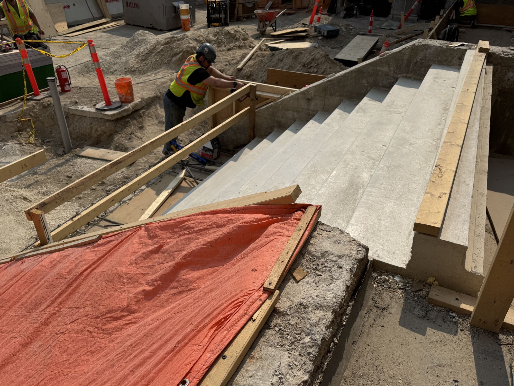 Looking down and to the right towards a set of poured concrete stairs leading up to the Rupert Ave entrance. The stairs are blocked at each end with plywood and construction fencing. In the lower left part of an orange tarpaulin can be seen covering the stairs going down to the Concourse entrance.