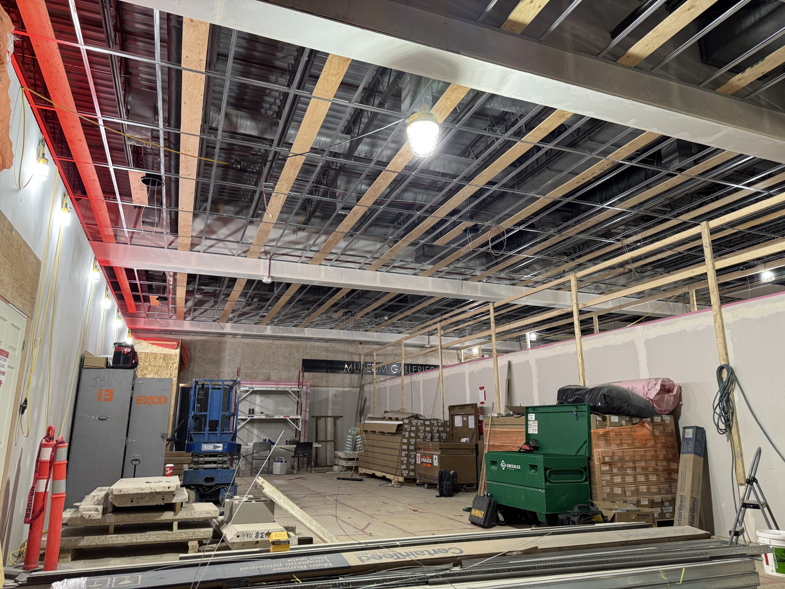 The north side of the Museum foyer under construction, with materials stacked about. The ceiling is uncovered, showing the pipes and vents, with original all light fixtures removed. New inter-com and light fixtures are being installed.