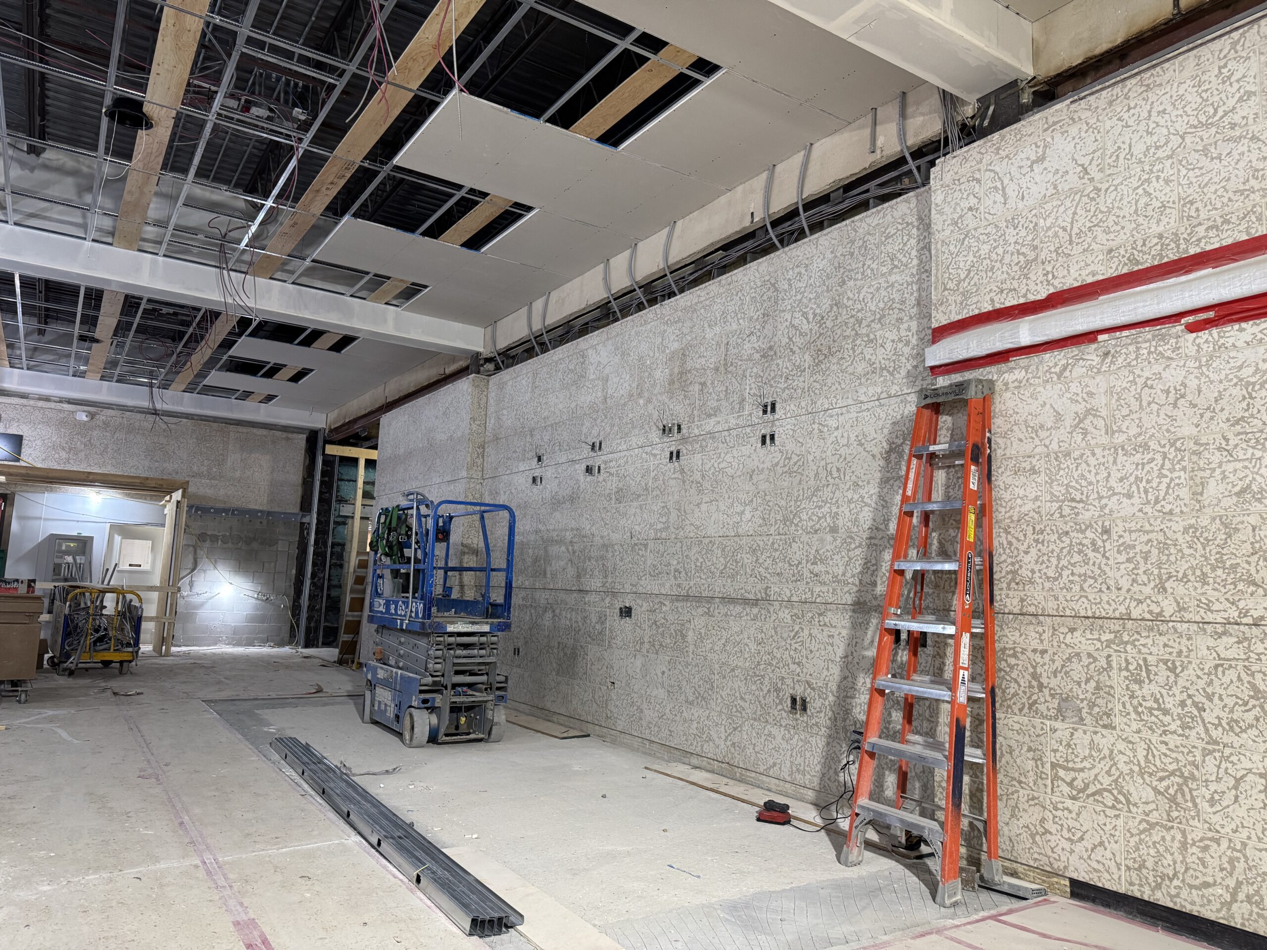 The south side of the museum foyer looking toward where the Box Office would be. Wiring is installed from teh Tyndall Stone on the back wall, and an orange ladder is folded back against the wall and a blue scissor lift is parked nearby. The ceiling is partial open, showing work on the lighting and intercom systems, and ceiling tiles are being replaced.