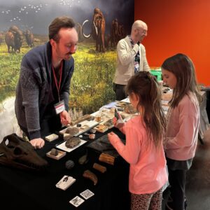 Two children stand at one side of a table displaying fossil specimens, with one of them holding a large tooth. On the other side of the table a Ed Leith Cretaceous Menagerie staff person talks with them.