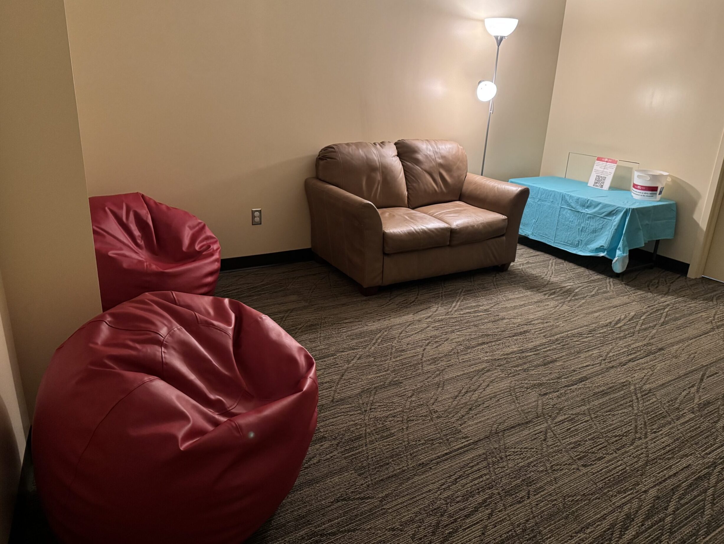A softly lit room with two red beanbag chairs and a beige two-seat couch next to a lamp and a low table with a blue tablecloth.