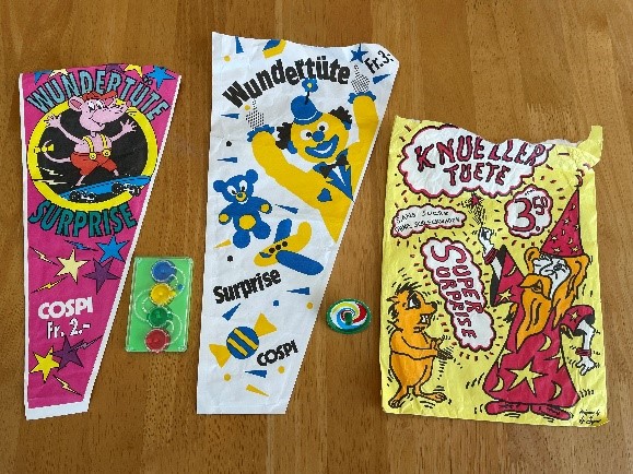Three empty paper bags decorated in bright colours with cartoon characters and text reading "Wundertute / Suprise / Cospi", with a small maze game and top between then.