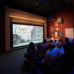 A group of people sit in benches facing a large video screen framed with stone accenting. On the wall to the right of the screen, a variety of artifacts are mounted on display.