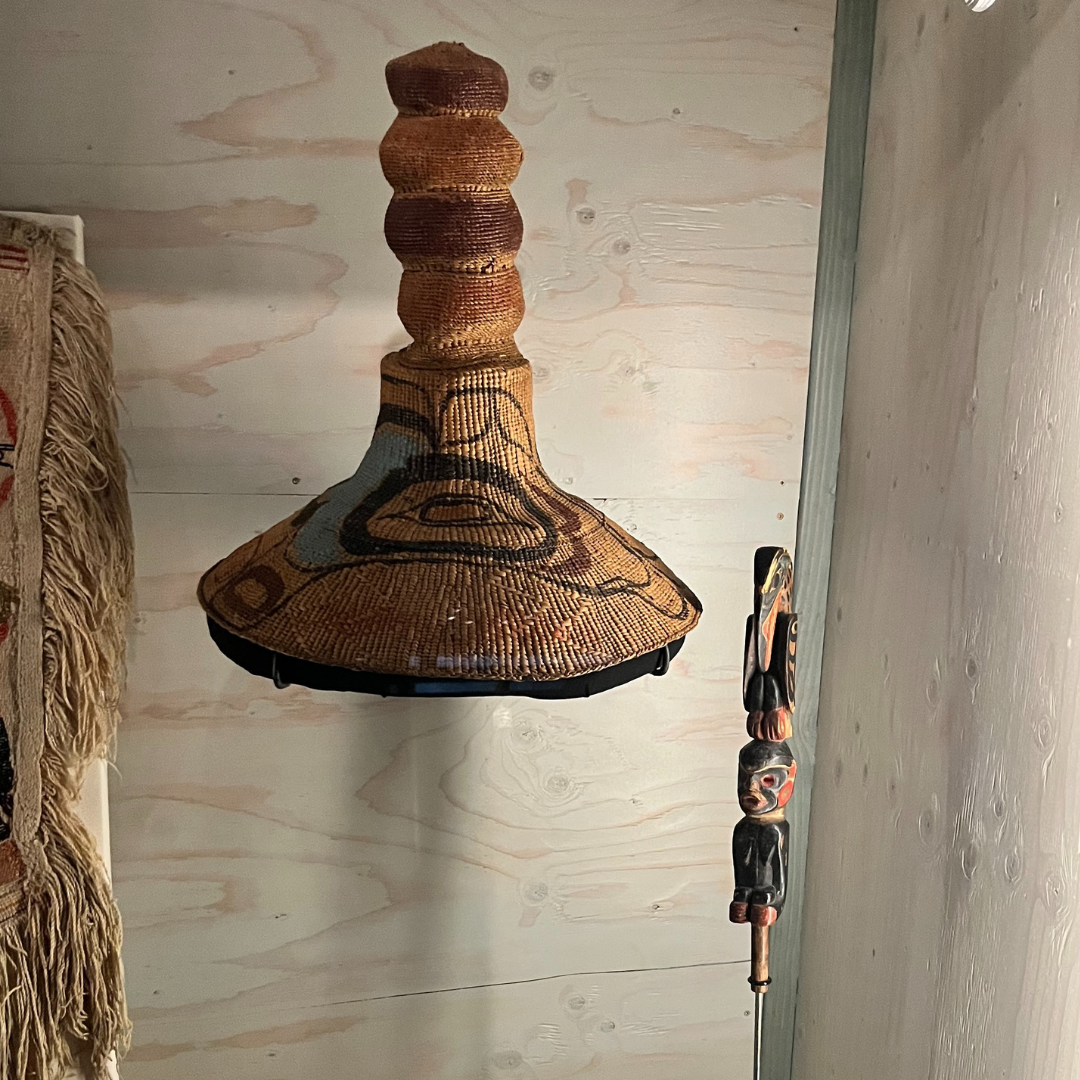 A hat on display in a museum case. The hat has a wide-brim and stacked circles coming up from the centre. Next to it is displayed a ornately carved staff.